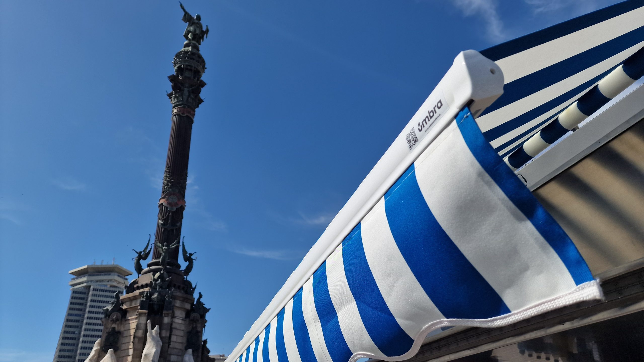 Toldo con rayas de color blanco y azul claro, en el fondo se ve la estátua de Colón de Barcelona