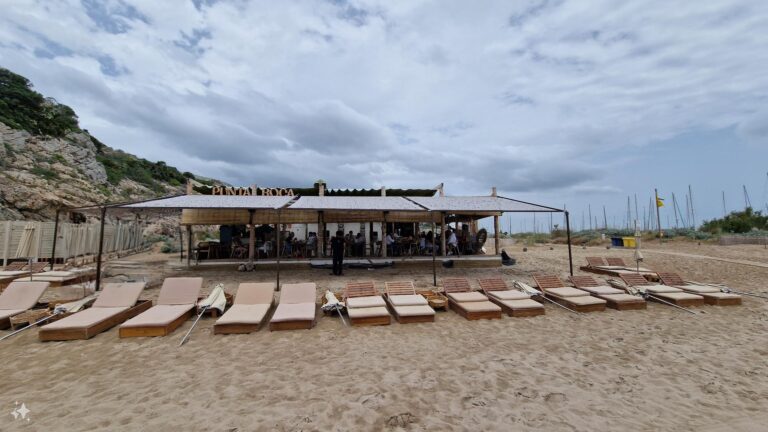Esta imagen muestra un restaurante de playa, "Punta Roca", con una amplia zona de terraza cubierta por un toldo claro y rodeada de tumbonas sobre la arena, creando un ambiente relajado junto al mar.