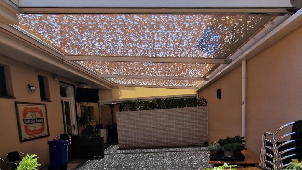 Vista de un patio o terraza parcialmente cubierto por una estructura de sombra tipo malla, proyectando un patrón de luz moteada sobre el suelo y el mobiliario.