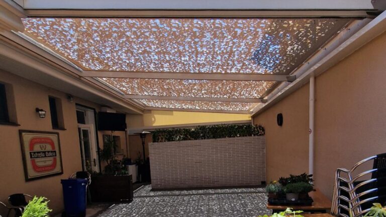 Vista de un patio o terraza parcialmente cubierto por una estructura de sombra tipo malla, proyectando un patrón de luz moteada sobre el suelo y el mobiliario.