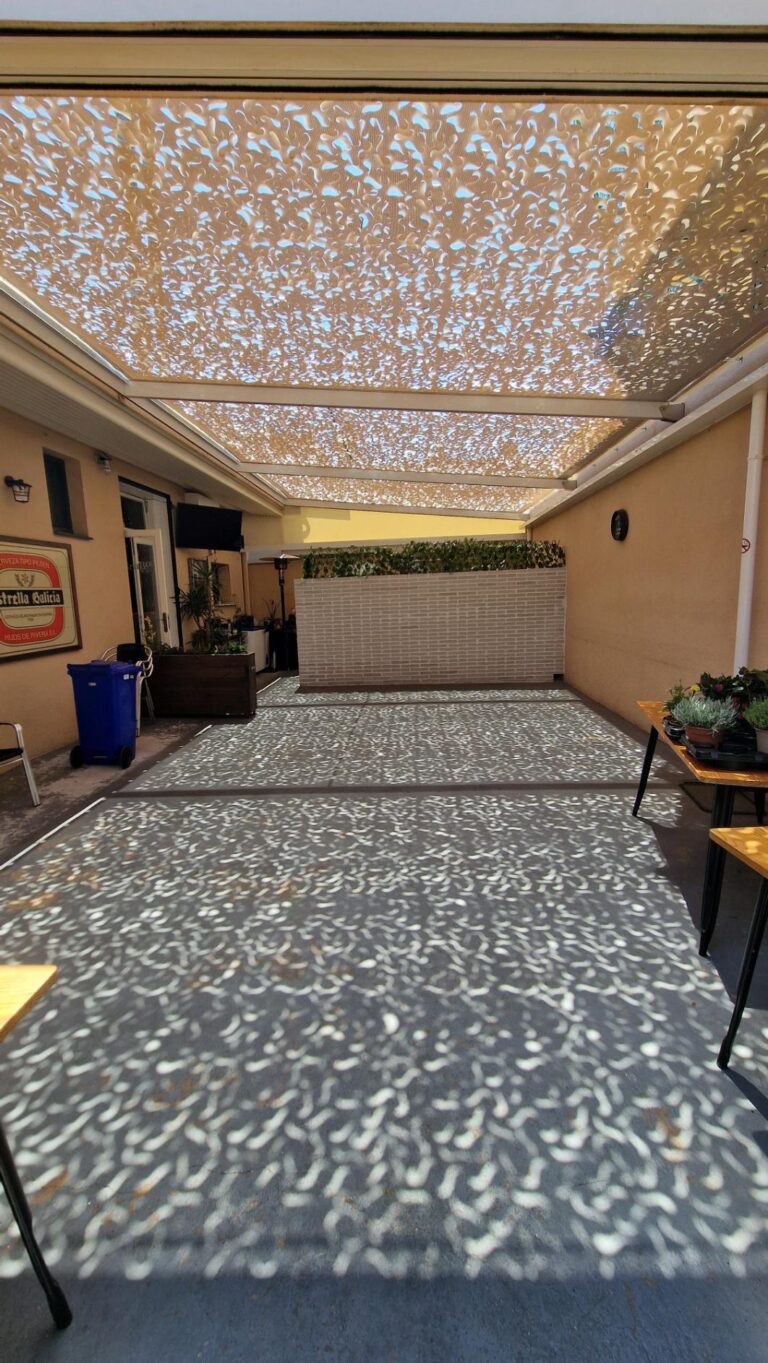 Vista de un patio o terraza parcialmente cubierto por una estructura de sombra tipo malla, proyectando un patrón de luz moteada sobre el suelo y el mobiliario.