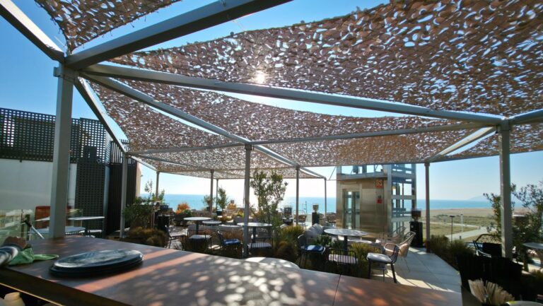 "Amplia terraza de un restaurante o bar con vistas al mar, cubierta por una estructura de sombreado tipo malla que crea patrones de luz y sombra sobre las mesas y el suelo, ofreciendo un ambiente exterior atractivo."