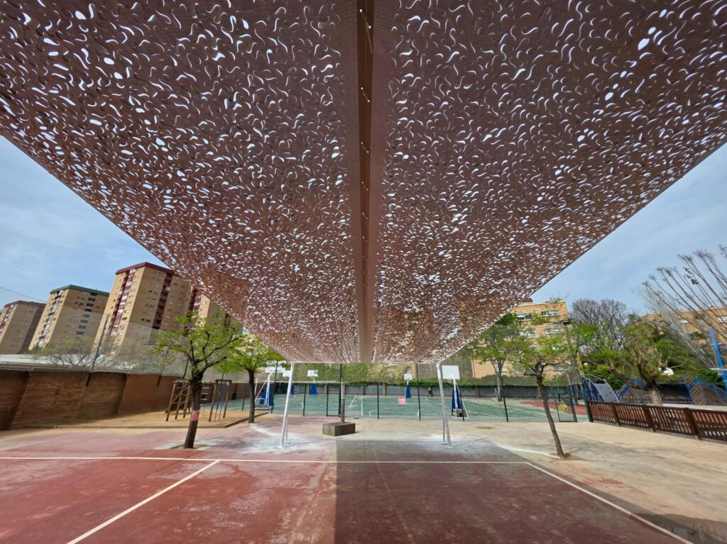 Tres velas de sombra de color beige, fijadas a mástiles, proyectan sombra sobre un patio de juegos escolar soleado con un tobogán y columpios.