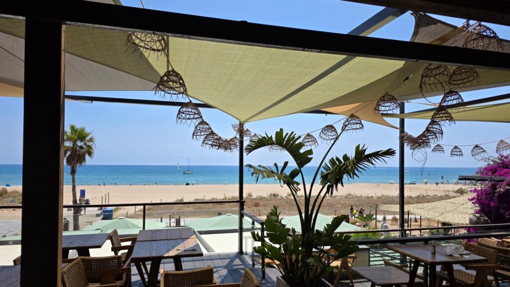 Una terraza de restaurante frente a la playa, con toldos que proporcionan sombra y decorada con luminarias colgantes de aspecto natural, ofreciendo vistas al mar.
