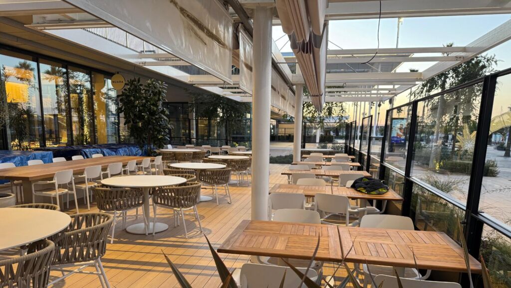 Terraza exterior de un restaurante con mesas y sillas de diseño moderno, protegida por un sistema de toldos o pérgolas retráctiles, y rodeada por paneles de vidrio y vegetación.