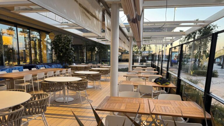 Terraza exterior de un restaurante con mesas y sillas de diseño moderno, protegida por un sistema de toldos o pérgolas retráctiles, y rodeada por paneles de vidrio y vegetación.