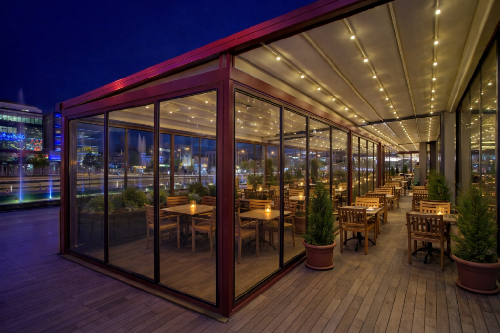 An empty outdoor restaurant terrace at night featuring a modern glass enclosure and a retractable fabric pergola roof with integrated LED strip lighting. The space is furnished with warm wooden tables and chairs, each set with a small glowing candle. Potted evergreen trees line the wooden deck, and blue-lit fountains are visible in the background against a deep blue twilight sky