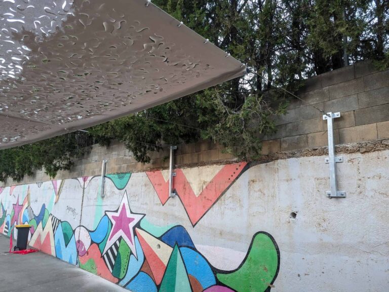 Vela de sombra calada anclada mediante cables y soportes metálicos a un muro con mural en la Escola Josep Trueta de Viladecans.
