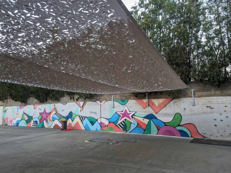 Vela de sombra calada anclada a muro sobre un patio escolar con mural multicolor en Viladecans.