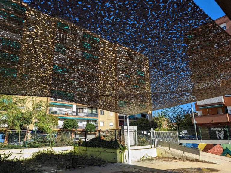 Vista inferior de malla de sombreo calada tipo camuflaje instalada sobre poste en el patio exterior de Escola Les Planes.