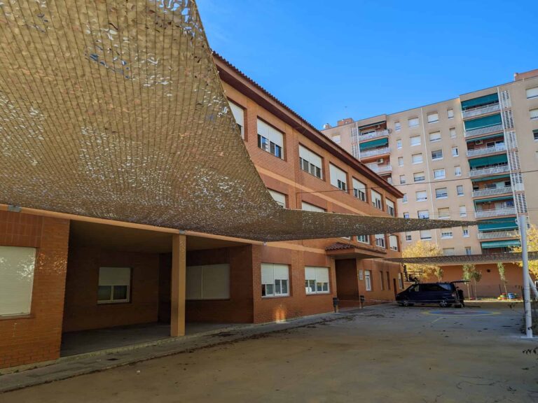 Vela de sombra calada instalada sobre el patio de la Escola Les Planes en La Llagosta, anclada a la fachada del edificio.