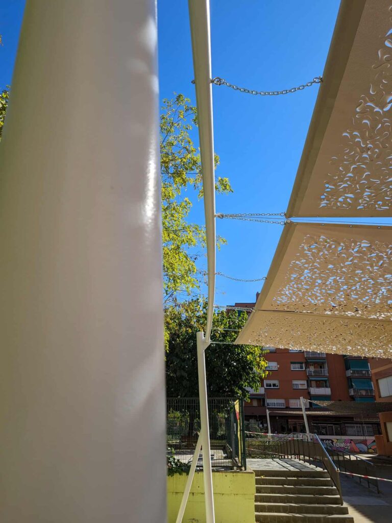 Detalle de poste blanco y fijaciones de una estructura de sombra con lonas caladas en el patio de Escola Les Planes.