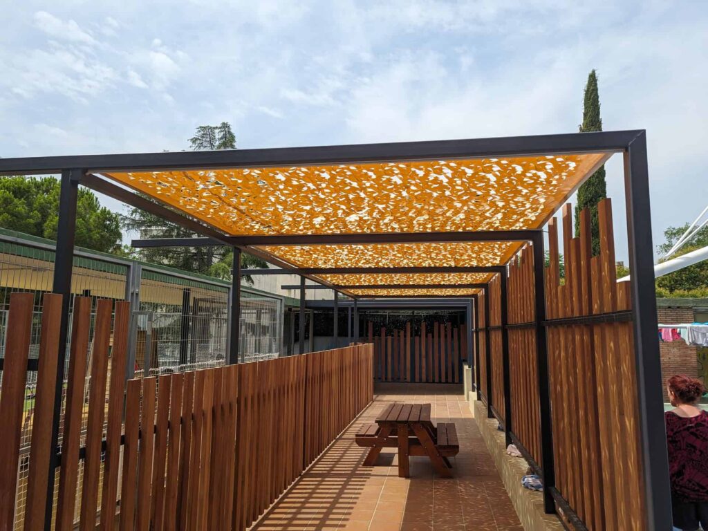 Pérgola con cubierta calada amarilla y cerramiento de madera en el patio de la Escola Regina Carmeli en Horta.