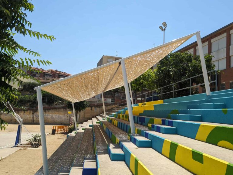 Estructura de sombra con tejido calado sobre las gradas pintadas del patio del Institut El Cairat en Esparreguera.