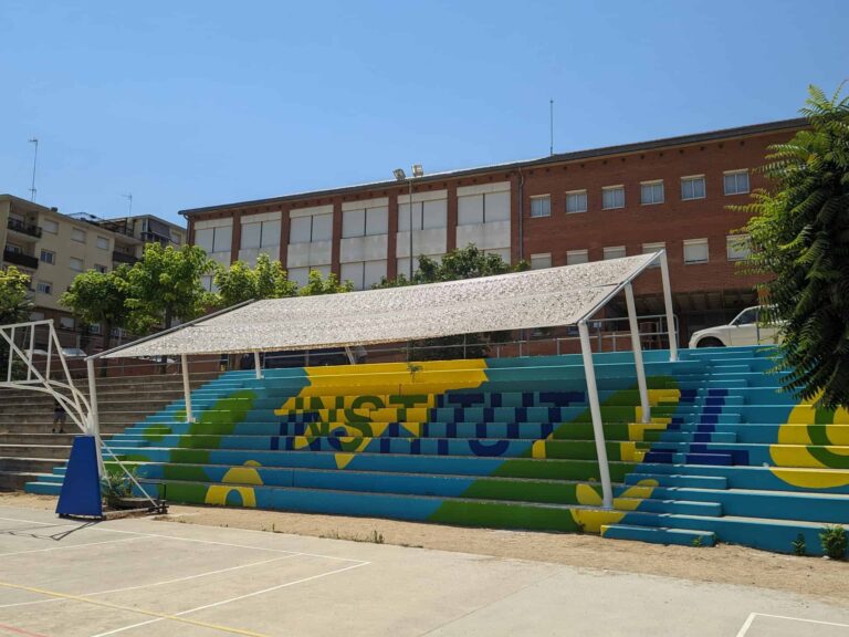 Estructura de sombra inclinada instalada sobre las gradas pintadas del patio del Institut El Cairat en Esparreguera.