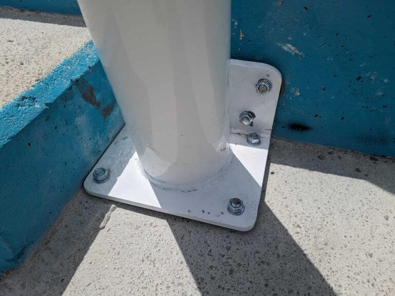 Detalle de fijación de poste blanco mediante placa de anclaje atornillada al suelo y murete en Institut El Cairat.