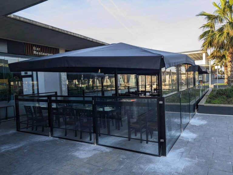 Terraza de restaurante cerrada con mamparas de cristal y estructura de sombra textil oscura en Port Premià.