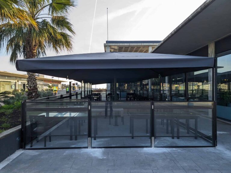 Terraza de restaurante con gran parasol oscuro y mamparas de cristal perimetrales en Port Premià.