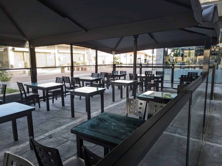 Terraza de restaurante con parasoles oscuros, mesas de comedor y cerramiento perimetral de mamparas de vidrio en Port Premià.