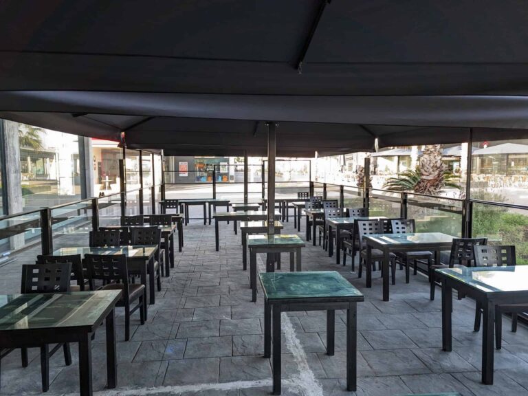 Terraza de restaurante con parasoles oscuros, mesas y mamparas perimetrales de cristal en Port Premià.