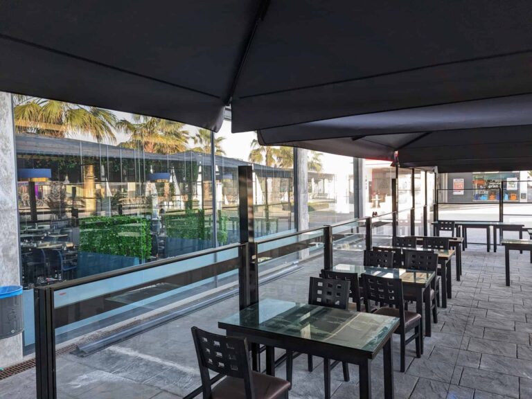 Terraza del restaurante Koi Sushi en Port Premià con parasoles oscuros, mamparas de cristal y mesas dispuestas en hilera.