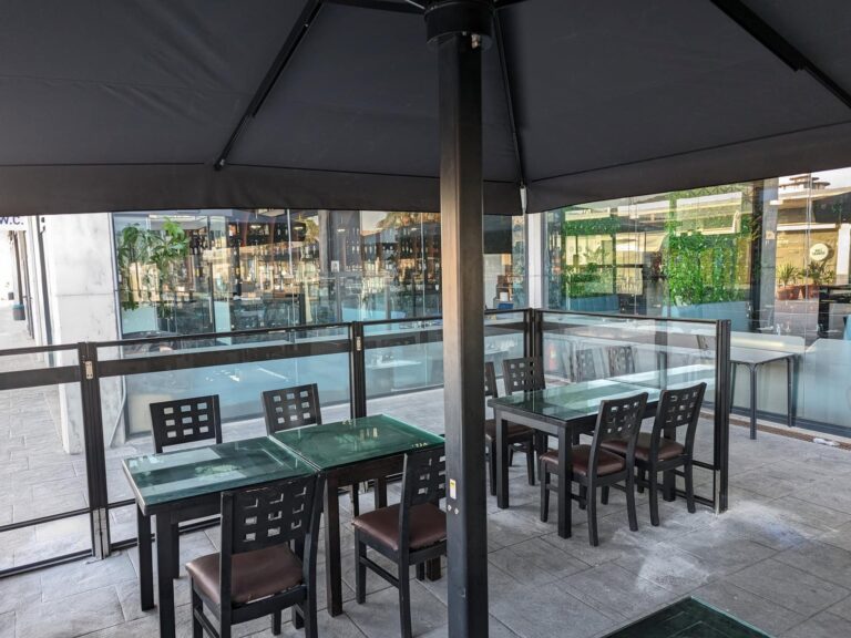 Terraza de restaurante protegida por parasol oscuro y mamparas de cristal con mesas de comedor exterior en Port Premià.
